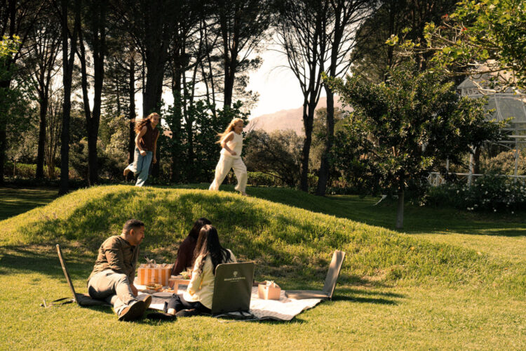 A family enjoying a lawn picnic on a blanket with low-back chairs while two young girls run barefoot across a grassy mound behind them under tall trees.