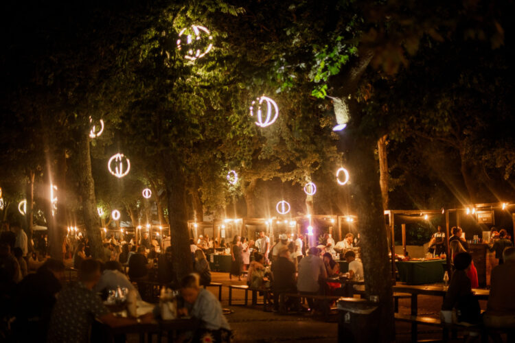Friday Night Market Crowds seated at long wooden trestle tables under oak trees at night, lit by glowing ring lights and string lights, with market stalls and the Boschendal Bar in the background.